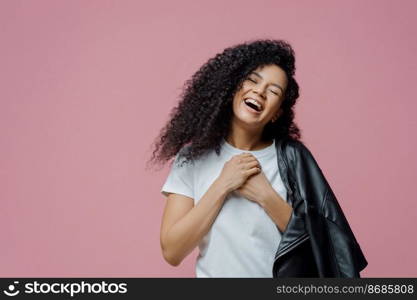 Overjoyed dark skinned woman keeps hands together and laughs sincerely, closes eyes and feels very happy, shows white perfect teeth, wears t shirt and leather jacket on shoulder, isolated on pink wall
