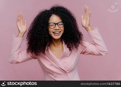 Overjoyed emotional female entrepreneur raises hands, laughs sincerely, wears spectacles and formal jacket, works in business sphere, poses indoor against purple background. Happy emotions concept