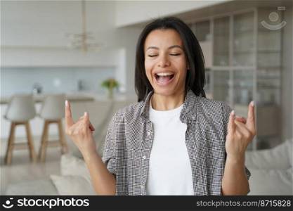 Overjoyed female showing hard rock hand gesture, standing at home. Excited happy cool young woman shows horns sign by fingers, screaming or singing favorite song, feeling success and festive.. Excited cool young woman shows hard rock hand gesture, screaming singing at home. Success, happiness