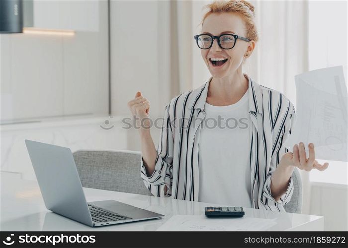 Overjoyed ginger woman holds papers works on project statistics celebrates finishing of work calculates finances household expenses sits at white table in front of opened laptop dressed casually. Overjoyed ginger woman holds papers works on project statistics celebrates finishing of work