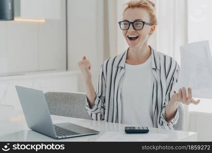 Overjoyed ginger woman holds papers works on project statistics celebrates finishing of work calculates finances household expenses sits at white table in front of opened laptop dressed casually. Overjoyed ginger woman holds papers works on project statistics celebrates finishing of work