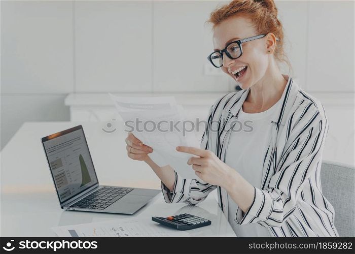 Overjoyed ginger woman reading reading financial paper document and smiling of pleasant news, receiving notification about last mortgage payment and feeling happy, calculating monthly budget at home. Overjoyed ginger woman reading reading financial paper document and smiling of pleasant news