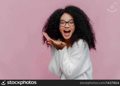 Overjoyed happy curly young woman with cheerful expression, keeps mouth widely opened, wears casual white jumper and optical glasses, poses over violet background. Emotions and happiness concept