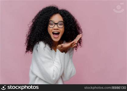 Overjoyed happy curly young woman with cheerful expression, keeps mouth widely opened, wears casual white jumper and optical glasses, poses over violet background. Emotions and happiness concept
