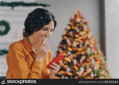 Overjoyed hispanic lady sits in living room decorated with Xmas tree looking at mobile screen with sweet smile while reading messages from friends or co-workers with Happy New Year and Merry Christmas. Overjoyed hispanic lady sits in living room decorated with Xmas tree looking at mobile screen