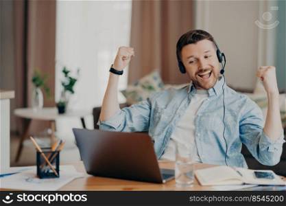 Overjoyed male freelancer in casual wear feeling excited and celebrating triumph after successful job interview online, wearing headset with microphone and sitting at his workplace at home. Handsome man enjoying victory while sitting at his workplace at home