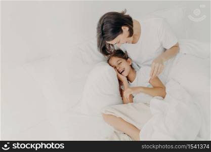 Overjoyed mum and daughter have fun together in bedroom, pose under soft blanket. Happy small girl spends leisure time with mother, has toothy smile, expresses happiness. Children and parents