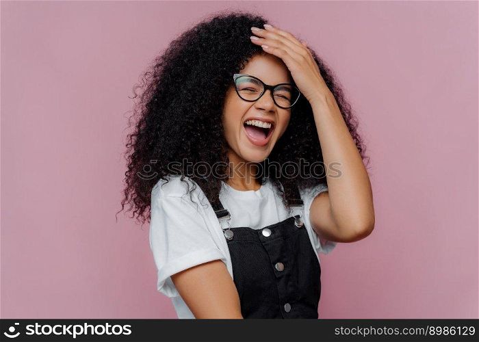 Overjoyed pleased curly haired woman keeps hand on forehead, laughs from happiness, understands she did something funny, wears optical glasses, casual outfit, models against purple background
