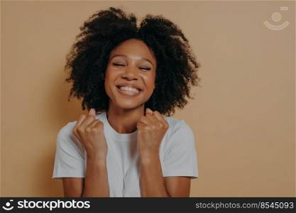Overjoyed pleased dark skinned young woman raises clenched fists, shows white teeth, wears casual white tshirt, models isolated over pastel beige background with copy space, celebrates excellent news. Overjoyed pleased happy dark skinned young woman raises clenched fists and smiling