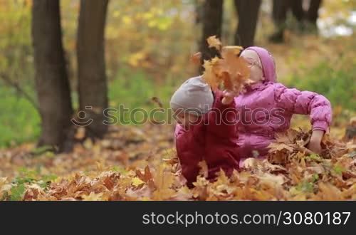 Overjoyed smiling siblings throwing yellow fallen maple leaves while playing together in pile of foliage in autumn park. Carefree cute teenage girl in eyeglasses and her toddler brother having fun outdoors in fall time. Slow motion.