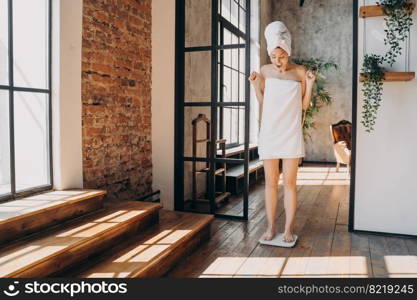 Overjoyed woman standing on floor scale, celebrating slimming success after shower at home. Happy female measuring weight after dieting or sports training. Healthy lifestyle, weightloss concept.. Excited woman standing on floor scale, measuring weight after dieting, celebrating slimming success