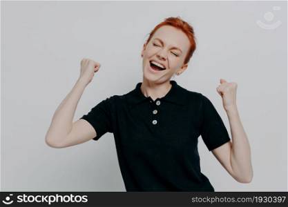Overjoyed young red haired female keeping eyes closed and screaming from excitement, raising clenched fists, celebrating success and triumph while stnading against grey wall, dressed in black t shirt. Overjoyed red haired female keeping eyes closed and screaming from excitement against grey wall