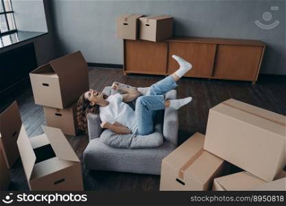 Overjoyed young woman has bought a new apartment. Happy hispanic lady relaxing in armchair. Pretty spanish girl at home with boxes. Concept of opportunity for woman. Independence and new lifestyle.. Overjoyed spanish young woman has bought a new apartment. Lady is relaxing in armchair.