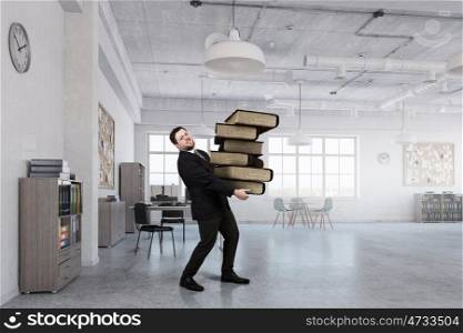 Overloaded with work. Young businessman in modern office carrying pile of books