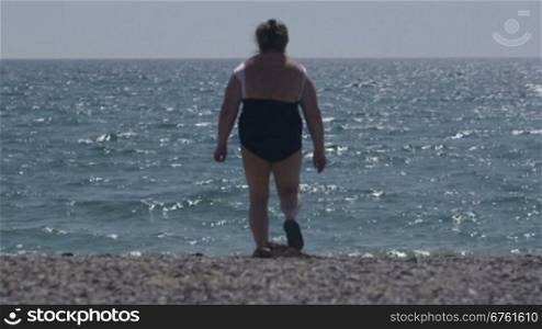 Overweight senior woman walking on the beach rear view