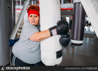 Overweight woman in gloves, boxing exercise with punching bag in gym. Calories burning, obese female person on hard training in sport club