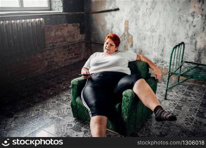 Overweight woman sits in a chair and watches TV. Unhealthy lifestyle, obesity
