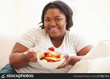 Overweight Woman Sitting On Sofa Eating Bowl Of Fresh Fruit