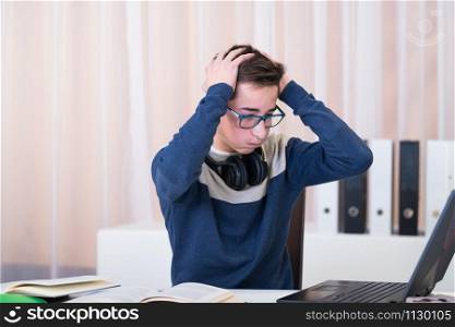 Overwhelmed teenage boy puts his hands to his head while watching a laptop screen on an out of focus background. Student concept.. Teenage boy puts his hands to his head
