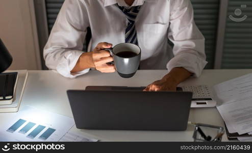 Overwork Concept An office man who works late drinking a cup of coffee while concentrating on his work on the notebook.