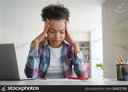 Overworked afro pupil is studying remote at home and getting headache. Teenage schoolgirl is sitting at the desk in front of laptop tired from homework. Problems with distance study at school.. Overworked pupil is studying remote at home and getting headache. Schoolgirl is tired from homework.