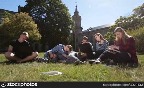 Overworked group of college students studiyng hard on university campus grass. Exhausted classmates learning together on park lawn while preparing for exams. Dolly shot.