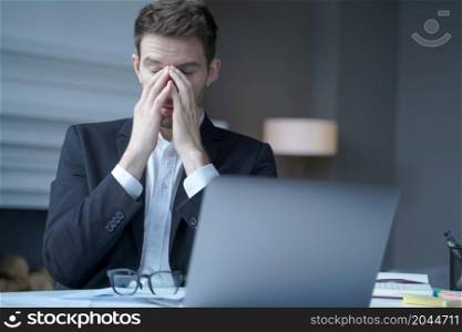 Overworked male entrepreneur sits at home office while took off glasses massages bridge of his nose with eyes closed, exhausted man suffering from heavy headache and fatigue. Health problem at work. Overworked male entrepreneur sits at home office, suffering from heavy headache and fatigue