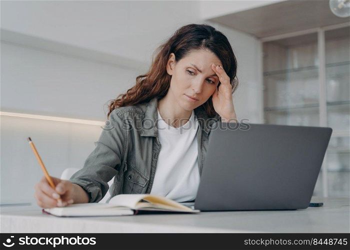 Overworked spanish lady has got a headache. Sick tired freelancer is working at home. Exhausted woman is chatting on laptop and taking notes with pencil. Frustrated businesswoman has problem.. Overworked spanish lady has got a headache. Sick tired freelancer is working on laptop at home.