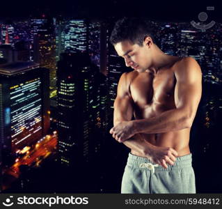 pain, sport, bodybuilding, health and people concept - young male bodybuilder touching injured elbow over night city background