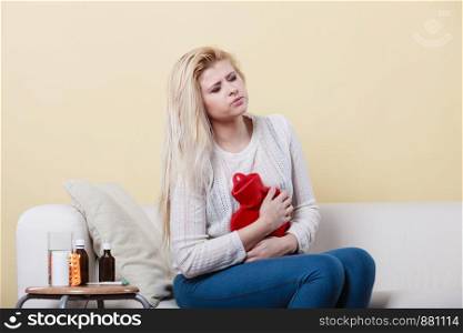 Painful periods and menstrual cramp problems concept. Woman having stomach cramps sitting on cofa feeling very unwell holding hot water bottle to feel some relief. Woman feeling stomach cramps sitting on cofa