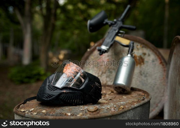 Paintball gun and protection mask closeup, nobody, playground in the forest on background. Extreme sport outdoors, pneumatic weapon and paint bullets, military team game concept. Paintball gun and protection mask closeup