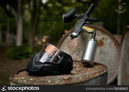 Paintball gun and protection mask closeup, nobody, playground in the forest on background. Extreme sport outdoors, pneumatic weapon and paint bullets, military team game concept. Paintball gun and protection mask closeup