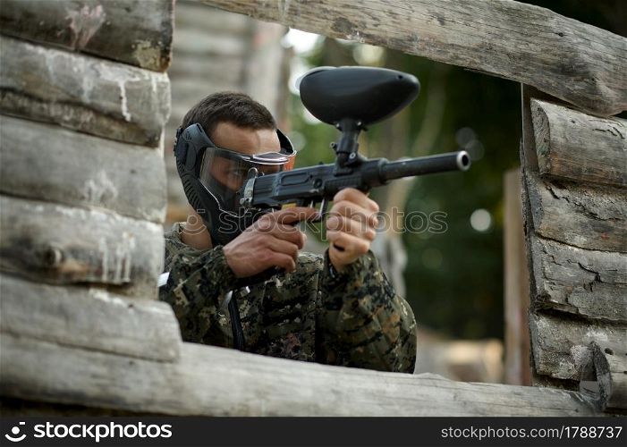 Paintball player aimimg with guns from the shelter on playground. Extreme sport with pneumatic weapon and paint bullets or markers, military game outdoors, combat tactics. Paintball player aimimg with guns from shelter