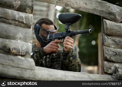 Paintball player aimimg with guns from the shelter on playground. Extreme sport with pneumatic weapon and paint bullets or markers, military game outdoors, combat tactics. Paintball player aimimg with guns from shelter