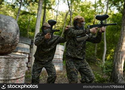 Paintball players in uniform and masks playing on playground in the forest. Extreme sport with pneumatic weapon and paint bullets or markers, military team game outdoors, combat tactics. Paintball players in uniform and masks in forest