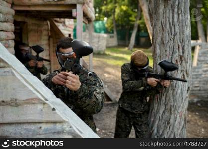 Paintball players in uniform and masks shoots with guns on playground in the forest. Extreme sport with pneumatic weapon and paint bullets or markers, military team game outdoors, combat tactics. Paintball players in uniform shoots with guns