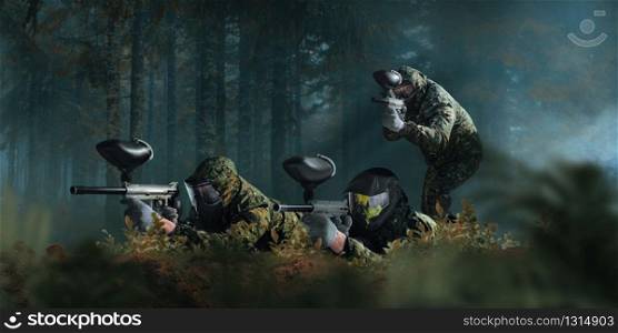 Paintball team shooting in forest battle. Extreme sport game, players in protection masks and camouflage lies on the ground and holds weapons in hands