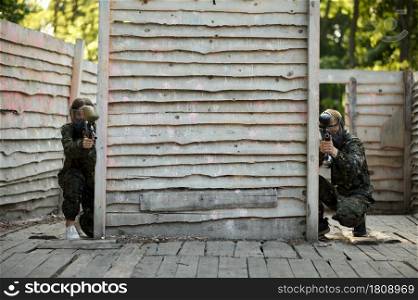 Paintball team shoots with guns from the shelter, warriors in camouflages on playground. Extreme sport with pneumatic weapon and paint bullets or markers, military game outdoors, combat tactics. Paintball team shoots with guns from the shelter