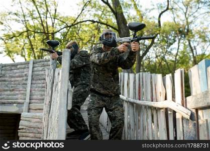 Paintball team shoots with guns, warriors in camouflages on playground in the forest. Extreme sport with pneumatic weapon and paint bullets or markers, active military game outdoors, combat tactics. Paintball team shoots with guns, military game