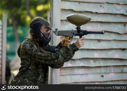 Paintball warrior shoots with guns from the shelter on playground. Extreme sport with pneumatic weapon and paint bullets or markers, military game outdoors, combat tactics. Paintball warrior shoots with gun from the shelter