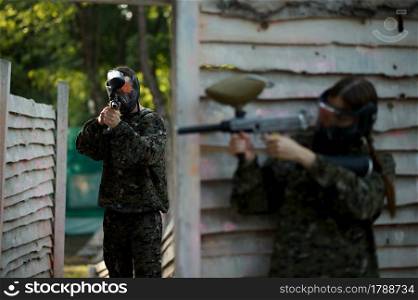 Paintball warrior shoots with guns from the shelter on playground. Extreme sport with pneumatic weapon and paint bullets or markers, military game outdoors, combat tactics. Paintball warrior shoots with gun from the shelter