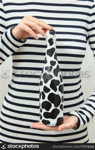 Painted black spots champagne bottle in woman&rsquo;s hands on striped shirt background, copy space.. Woman dressed in striped shirt holds in hands painted spots wine bottle.