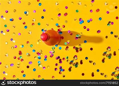 Painted red bottle on an yellow background covered colorful confetti with hard shadows, copy space. Holiday greeting card.. Holiday background with red painted bottle and colored confetti.