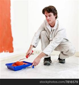 Painter, kneeling down to refill his paint roller with orange paint, whilst redecorating a room