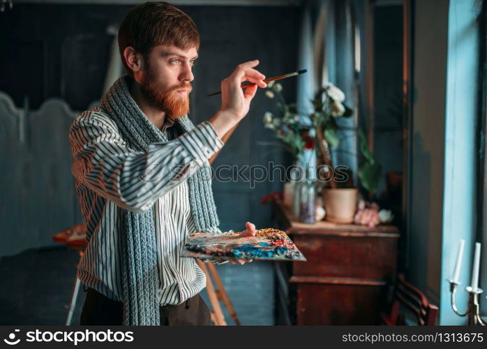 Painter with palette and brush in hand selective focus, art studio on background. Oil paint, paintbrush drawing. Painter with brush in hand selective focus