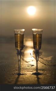 Pair of flute glasses filled with champagne with bows on stems on beach at sunset.