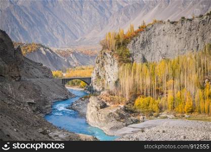 Pakistan in autumn. Clear turquoise blue river flow under the Ganish bridge between Hunza valley and Nagar, surrounded by colorful trees and mountains near Karakoram highway. Gilgit Baltistan.