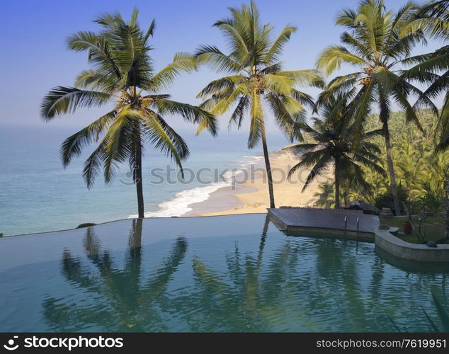 Palm trees are reflected in the pool and the ocean in a background. India.