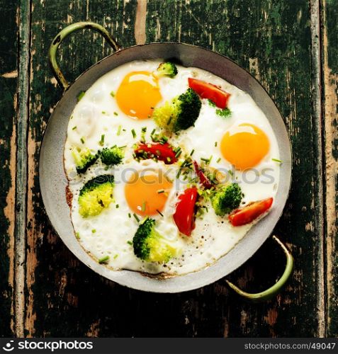 Pan of fried eggs, broccoli and cherry-tomatoes on old green background, top view