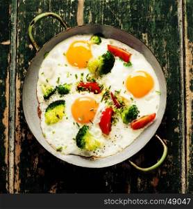 Pan of fried eggs, broccoli and cherry-tomatoes on old green background, top view